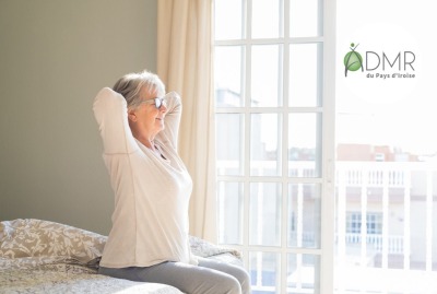 femme senior s'étirant devant une fenêtre, sur le bord de son lit à son réveil