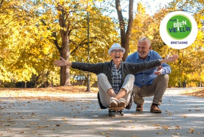 femme senior assise sur un skate qui roule pendant qu'un ami la regarde en souriant